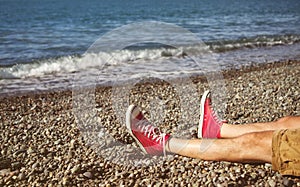 Men`s legs in shorts and red sneakers on a pebbly beach by the sea on a sunny day.