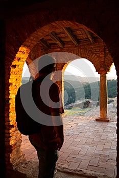 A men`s silhouette with a backpack stands in the opening of the arch of an old red brick building illuminated by the sunset.