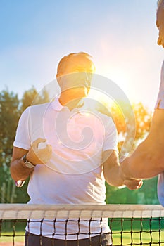Men shaking hands while standing by tennis net against clear sky on sunny day