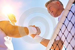 Men shaking hands while standing by tennis net against clear sky on sunny day