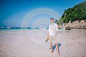 A men stands on a white sandy beach by the ocean. Men is holding a pineapple in his hand.