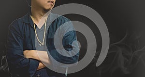 Men wear gold necklace in blue shirt stand in front of the smoke with light black background.