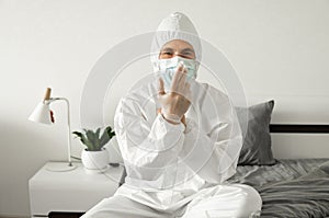 Men in white protective suit, medical mask and rubber gloves is sitting on a bed in his bedroom while coronavirus