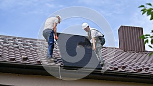 Men workers lifting up solar panel with help of ropes on roof of house.
