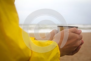 Men in yellow raincoat on the beach over the stormy sea holding a cup of hot tea