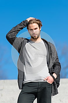 Mens fashion and style. Man with beard in casual wear touch hair on sunny outdoor, fashion. Macho stand on blue sky, perspective.