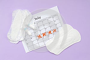 Menstrual pads and tampons on menstruation period calendar flat lay on lilac background