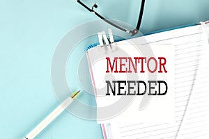 MENTOR NEEDED text on a sticky on notebook with pen and glasses , blue background