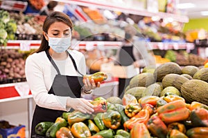 Merchandiser in protective mask lays out ripe bell peppers on the shelves in supermarket