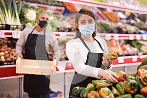 Merchandiser in protective mask lays out ripe bell peppers on the shelves in supermarket