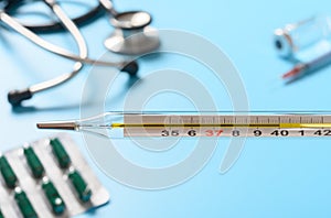 Mercury thermometer at 40 degrees centigrade with syringe and vaccine and stethoscope and capsules on the background
