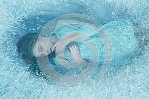 A mermaid girl in a blue vintage dress lies at the bottom of the lake. It is covered with ice edge, fish swim around it