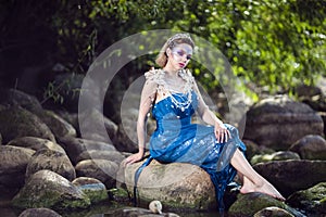 Mermaid of Sea in Artistic Caucasian Blond Woman With Strasses on Face Sitting in Blue Wet Dress on Rocky Shore Outdoor