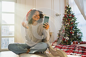 Merry Christmas. African woman in Santa hat having video chat in smartphone. Blogger in festive mood recording video in