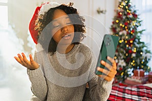 Merry Christmas. African woman in Santa hat having video chat in smartphone. Blogger in festive mood recording video in