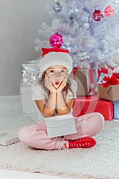 Merry Christmas celebration. Cheerful beautiful little girl sitting near the Christmas tree holding a gift box