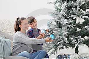 Merry christmas family at home, smiling brother and sister decorate the tree hang blue balls sitting on the floor in livong room