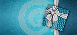 Merry christmas gift card, blue box with shiny silver ribbon bow isolated on turquoise glittering background, top view copy space