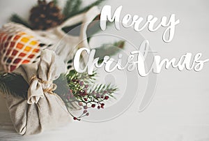 Merry Christmas Greeting card. Merry Christmas text handwritten on stylish furoshiki christmas gifts on rustic table with fir,