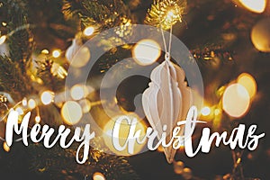 Merry Christmas greeting card. Merry Christmas text on modern christmas tree white bauble, golden christmas lights bokeh. Happy