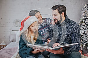 Merry Christmas and Happy Holidays Cheerful mom, dad and her cute daughter girl reading a book. Parent and little child