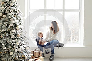 Merry Christmas and Happy Holidays! Cheerful mom and her cute baby boy with gift box near window and Christmas tree indoors