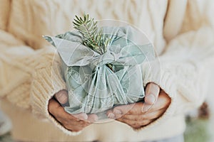 Merry Christmas and Happy Holidays! Hands holding gift wrapped in green fabric with fir branch. Stylish Furoshiki gift. Woman in