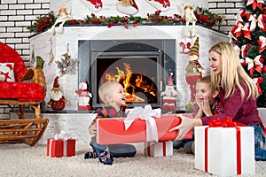 Merry Christmas and Happy Holidays!Mother and sons sitting by the fireplace and open Christmas gifts from Santa.