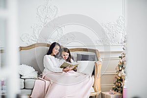 Merry Christmas and Happy Holidays. Pretty young mom reading a book to her cute daughter near Christmas tree indoors