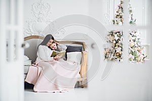 Merry Christmas and Happy Holidays. Pretty young mom reading a book to her cute daughter near Christmas tree indoors