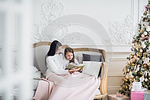 Merry Christmas and Happy Holidays. Pretty young mom reading a book to her cute daughter near Christmas tree indoors