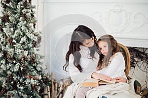 Merry Christmas and Happy Holidays, Pretty young mom reading a book to her cute daughter near tree indoors.