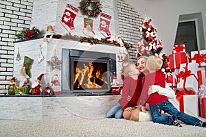 Merry Christmas and Happy Holidays!Two little sons kiss mom. The family is resting under a Christmas tree by the fireplace.