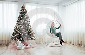 Merry Christmas and Happy New Year. Beautiful blonde woman in a green dress sitting in chair suspended at the tree