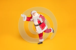 Merry Christmas and a happy new year ! Real Santa Claus carrying heavy sack with gifts, running fast on yellow studio background