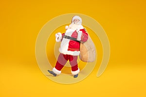 Merry Christmas and a happy new year ! Real Santa Claus carrying heavy sack with gifts, standing on yellow studio background .