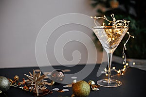 Merry Christmas and Happy New Year! Stylish christmas lights in champagne glass, golden confetti and baubles on moody background.