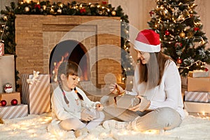 Merry Christmas! Mother and child girl with gifts at home, sitting on floor with crossed legs, being excited and happy, looking at