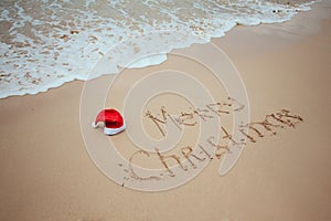 Merry Christmas with Santa red hat written on sand by hand on the tropical beach with ocean. New Year celebration on vacation