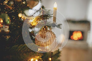 Merry Christmas! Stylish christmas gold bauble on tree close up against burning fireplace. Beautiful decorated christmas tree with