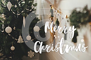 Merry Christmas text on stylish boho christmas tree, white baubles, garland, rustic stocking, lights. Season`s greetings card.