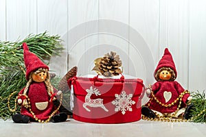 Merry New Year mood. Dolls, New Year`s decor, branches of a Christmas tree and a gift in a red round box on a light wooden.