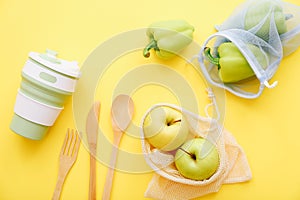 Mesh shopping bags with apples and peppers with reusable cup and wooden cutlery on yellow background. Top view. Eco