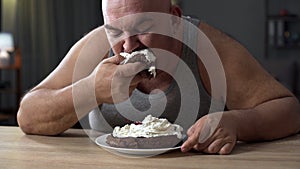 Messy obese man greedily eating cake with whipped cream, addiction to sweets
