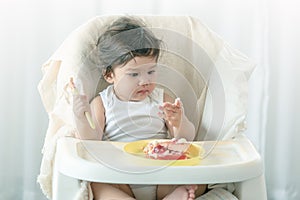 Messy one year baby girl eating cake.  Parents leave lovely infant girl holding a spoon, enjoying eating cake. Curious baby learns