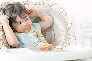 Messy one year baby girl eating pasta.  Parents leave lovely infant girl, enjoying eating meal alone. Curious baby learns eating