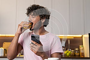 A mestizo guy stands in profile in the kitchen in subdued lighting, holds a phone and hamburger in hands, seductively