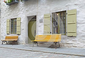 Metal benches in yellow. Facade of house with sash windows in green. White grainy wall Cobbled street. Canada