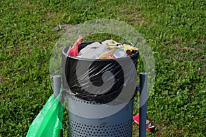 A metal bin full of rubbish, with a green plastic bag in a meadow photo