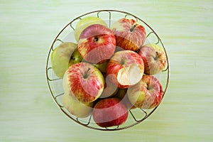 Metal bowl with green, yellow and red apples and one bitten apple on the green wooden background.